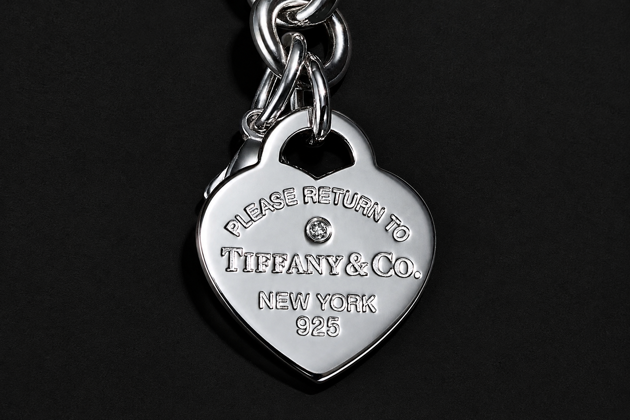 Silver heart tag reading Please Return To Tiffany & Co. New York 925, hanging from a chain on black.